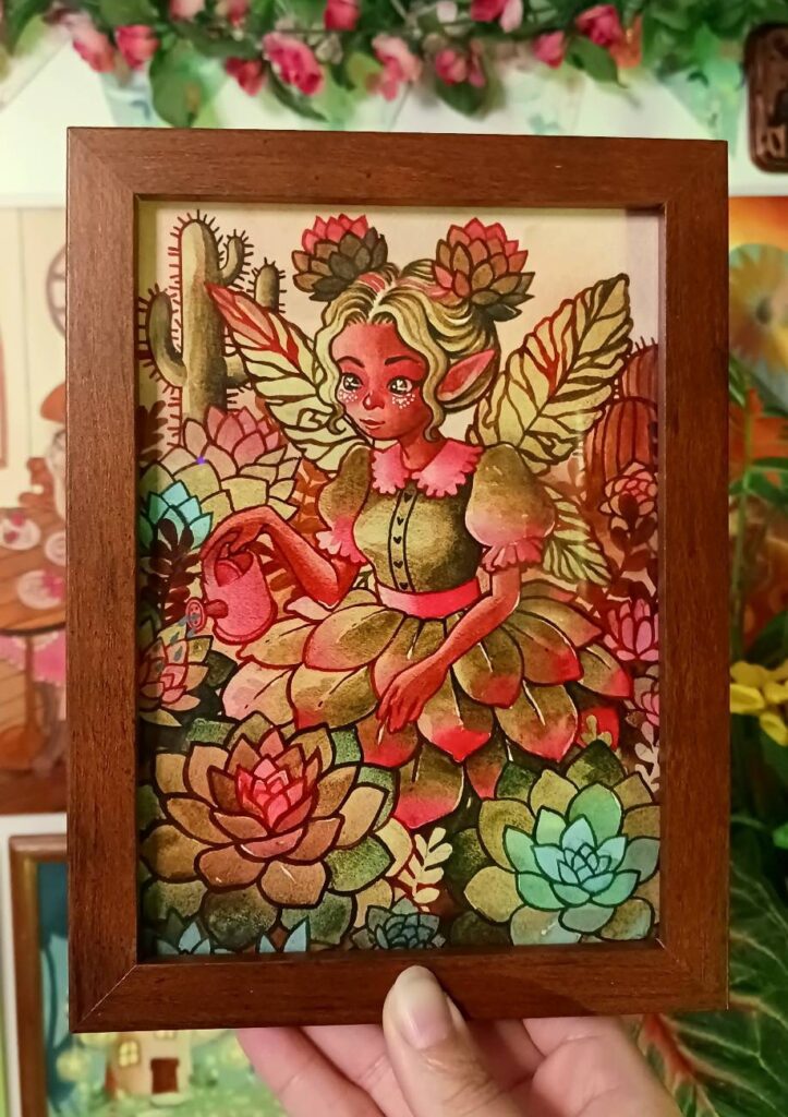 Framed fantasy artwork of a succulent themed fairy surrounded by colorful succulents and cacti. The fairy has wings, pointed ears, and a green and red petal dress, holding a pink watering can  in a vibrant botanical garden scene. Wooden frame held in a hand, promoting art prints available in an Etsy shop.
