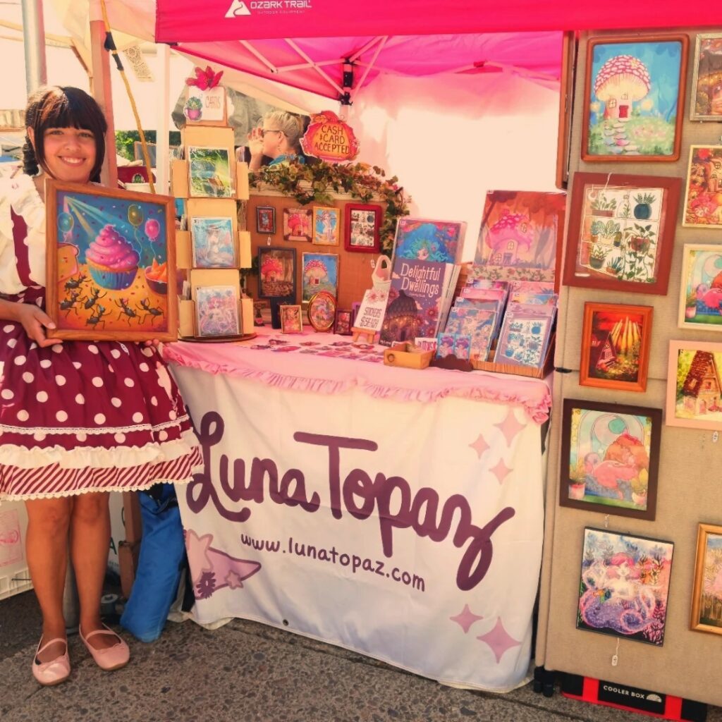 Artist Luna Topaz standing in front of a colorful craft market booth displaying whimsical fantasy artwork, framed prints, and illustrations under a red canopy.